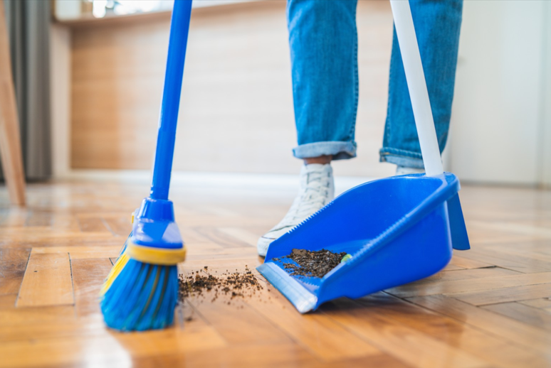 A person using a broom and a dustpan sweeping dirt off the wood floor. 